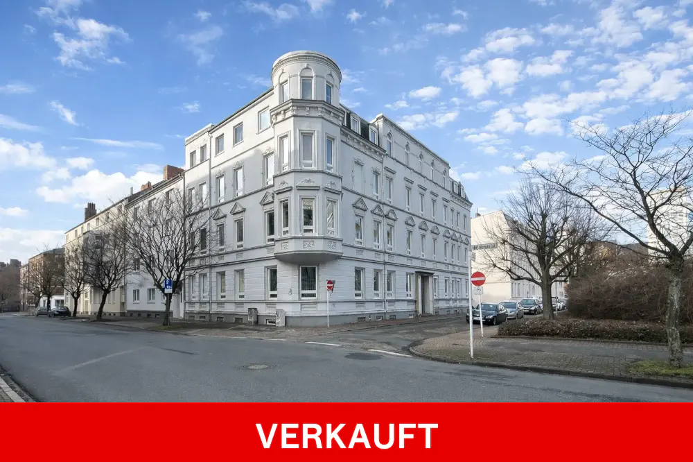 A light gray, multi-story building with a rounded corner tower, set on a city street under a cloudy sky. "VERKAUFT" is written in white on a red banner.