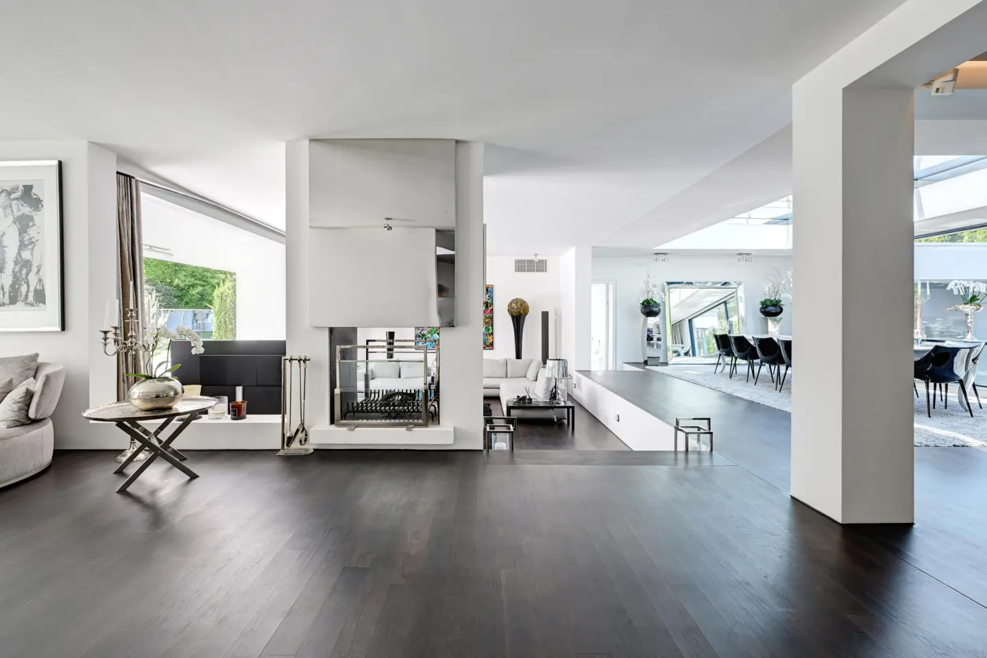 Open-concept living room with dark wood floors, white walls, and a modern fireplace. A dining area is visible in the background.