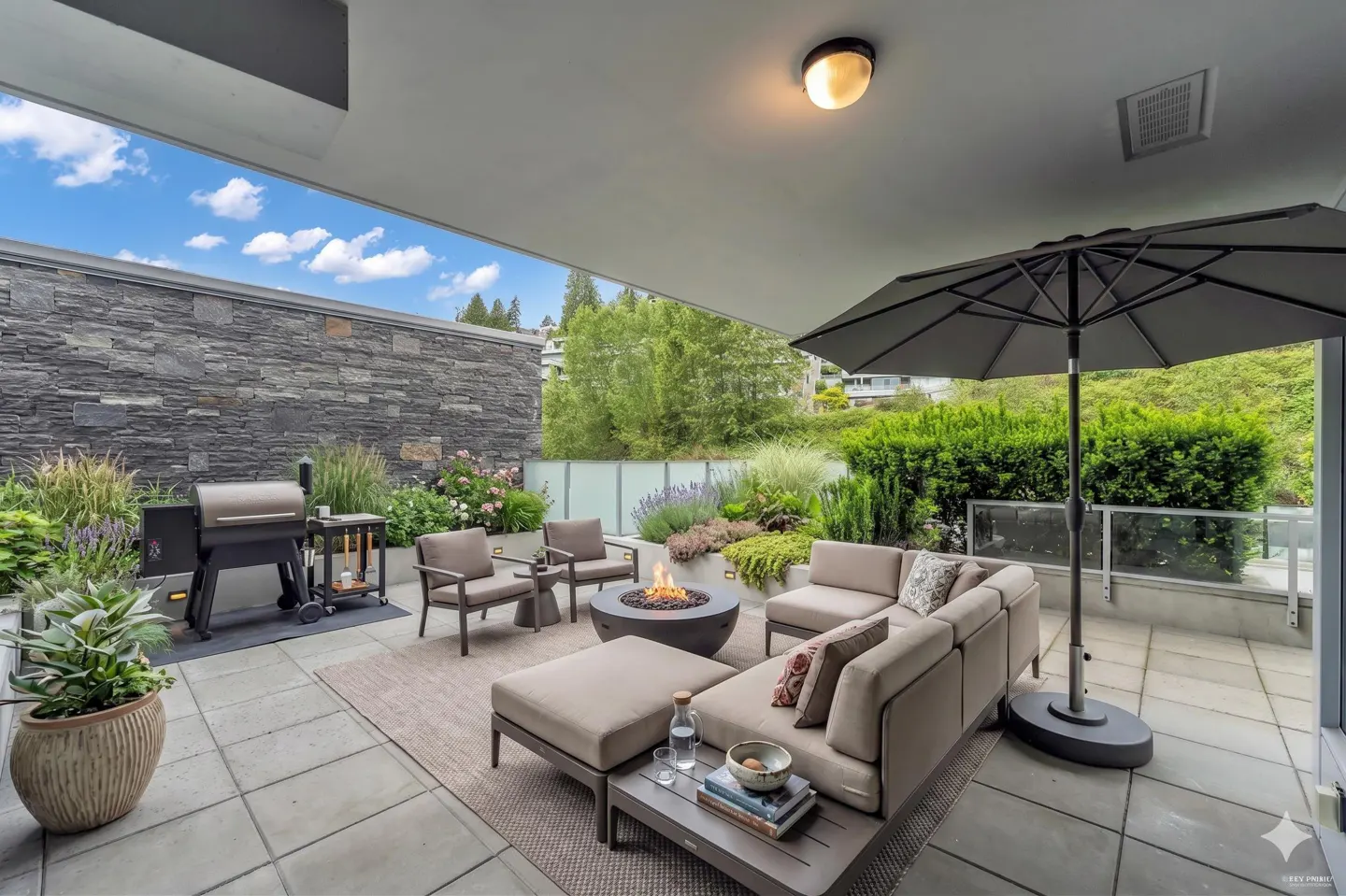 Outdoor patio with stone wall, grill, and seating area. A fire pit sits in the center, surrounded by chairs and a sectional sofa under an umbrella.