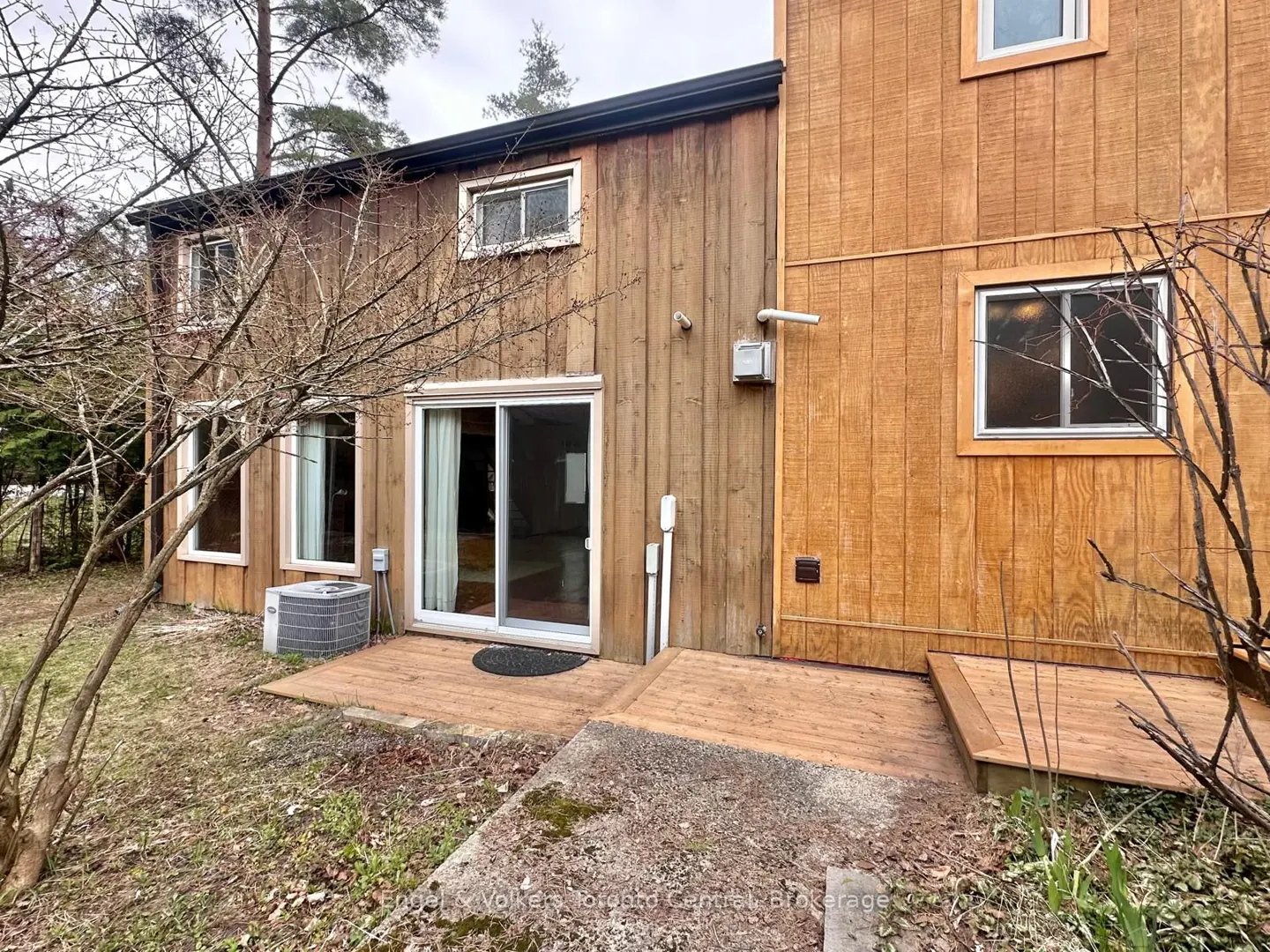 Exterior view of a two-story wood-sided house with a sliding glass door leading to a wooden deck and a concrete patio.