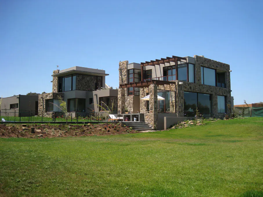 Two modern homes with stone and gray siding, large windows, and green lawn under a blue sky.