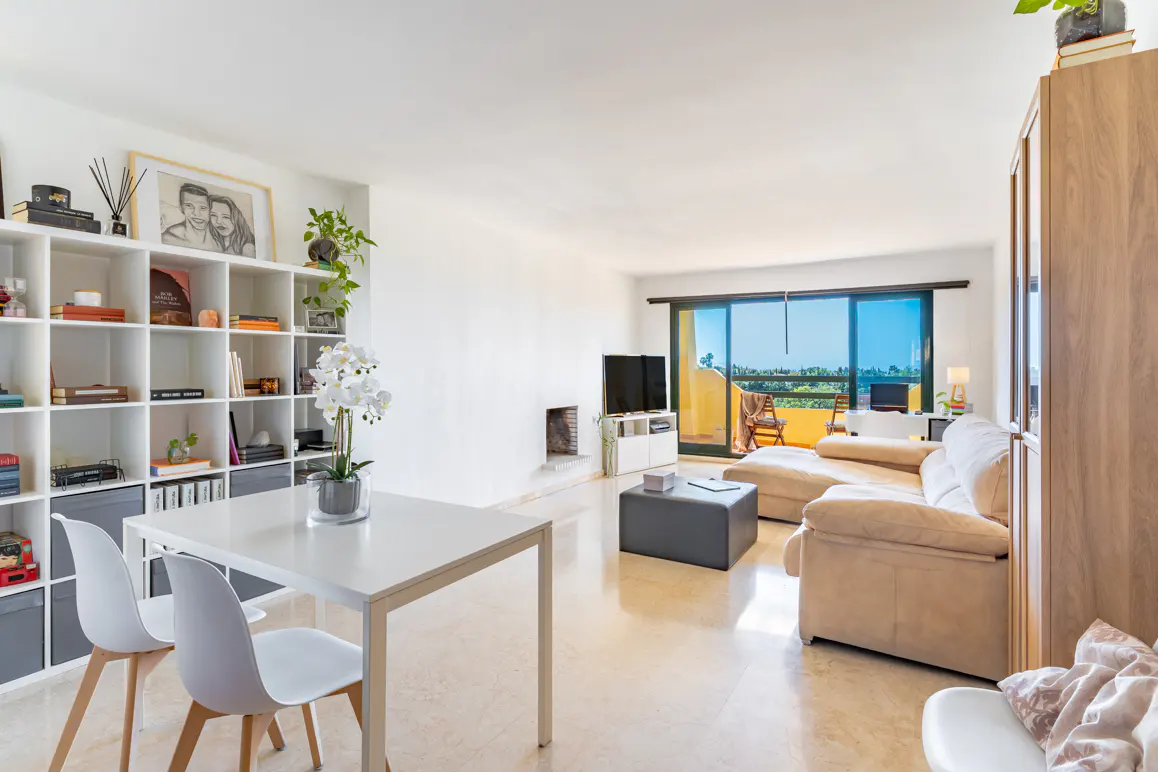 Bright living room with white walls, a beige sofa, and a white table with two chairs. A bookshelf is filled with books and decor. Balcony with a view.