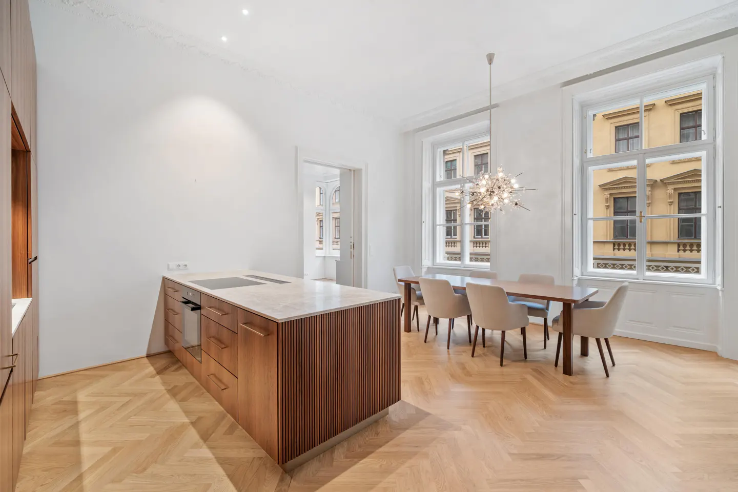 Bright, modern kitchen with wood floors, island, and dining table. Large windows offer a view of a building across the street.
