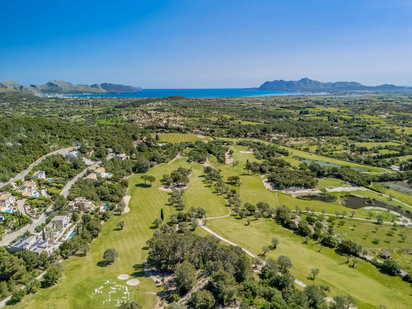 Aerial view of a green golf course with trees, houses, and mountains in the background under a clear blue sky.