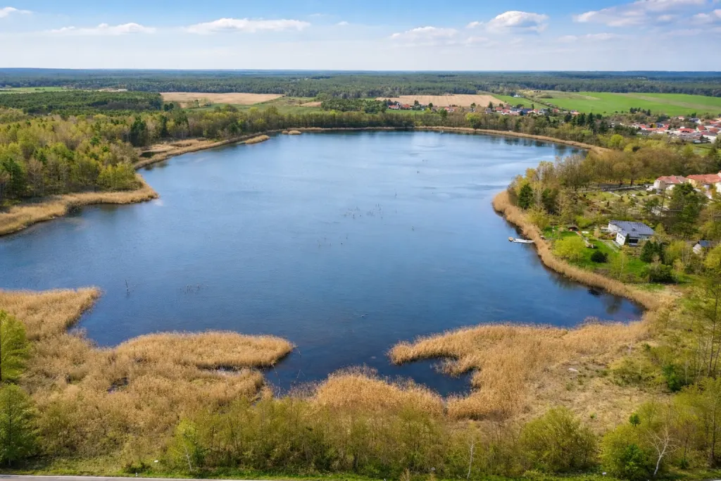 Aerial view of a blue lake surrounded by reeds, trees, and houses under a partly cloudy sky.