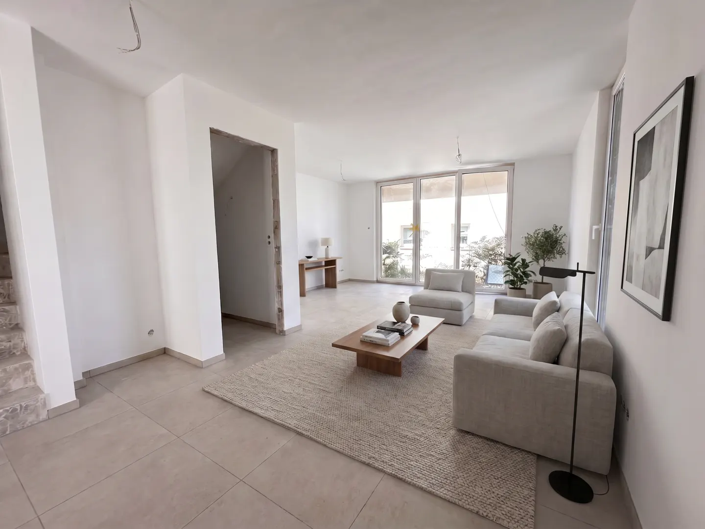 Bright, open living room with white walls, beige tile floor, and a large beige rug. A sofa, chair, and coffee table furnish the space. Large windows let in natural light.