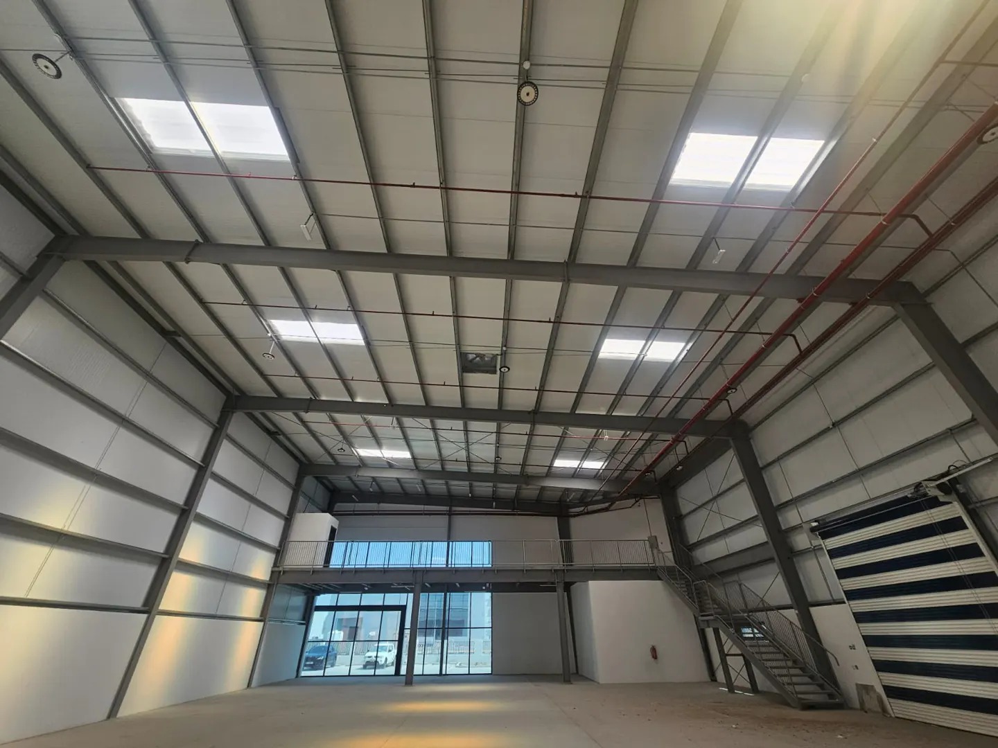 Interior of a large, empty warehouse with metal beams, skylights, and a mezzanine level with glass windows.