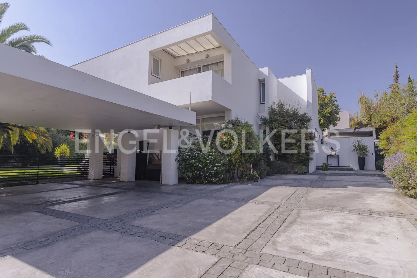 Modern white two-story house with a carport and a paved driveway. Lush greenery surrounds the property.