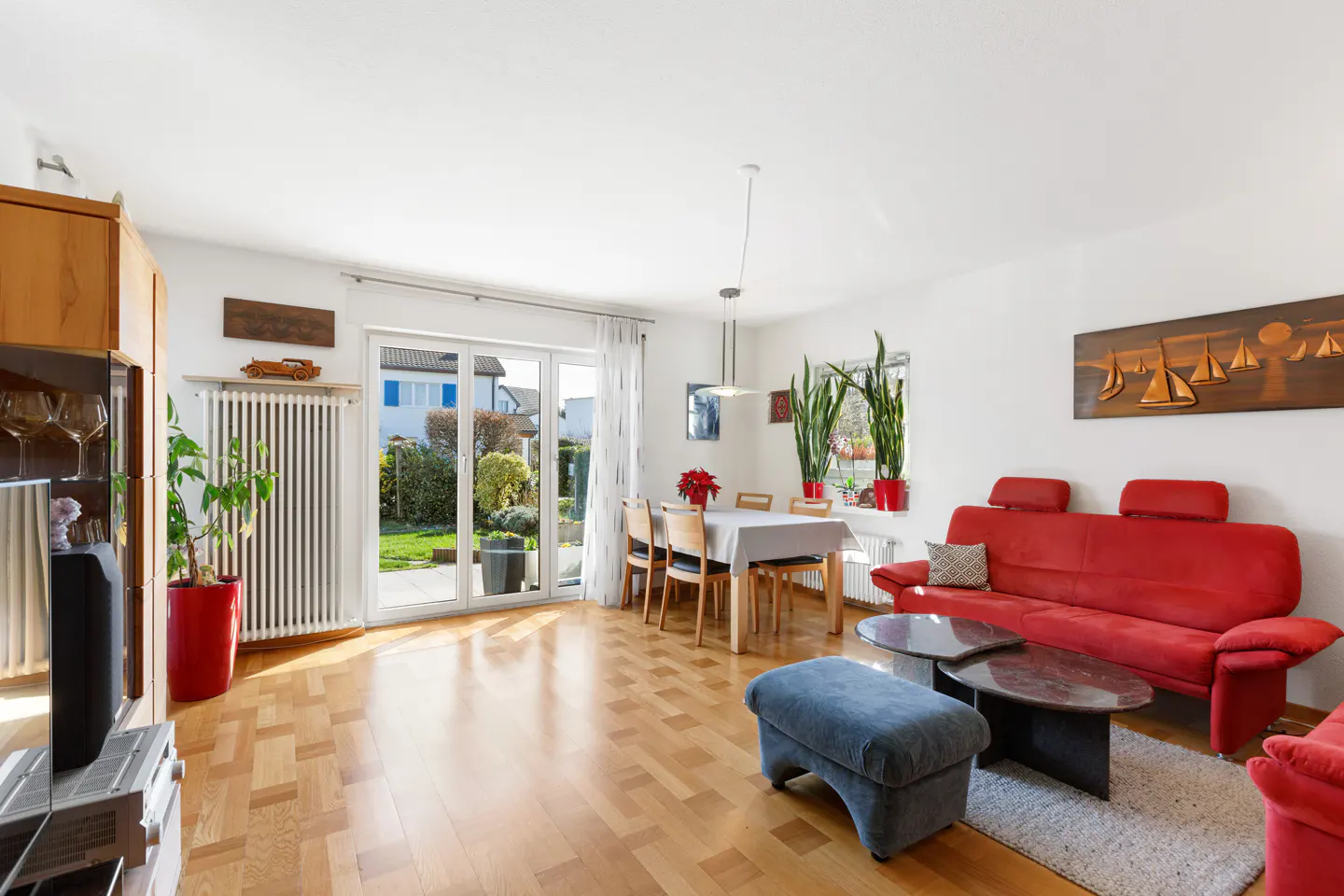 Bright living room with wood floors, red sofa, blue ottoman, and dining table. Patio doors lead to a green garden.