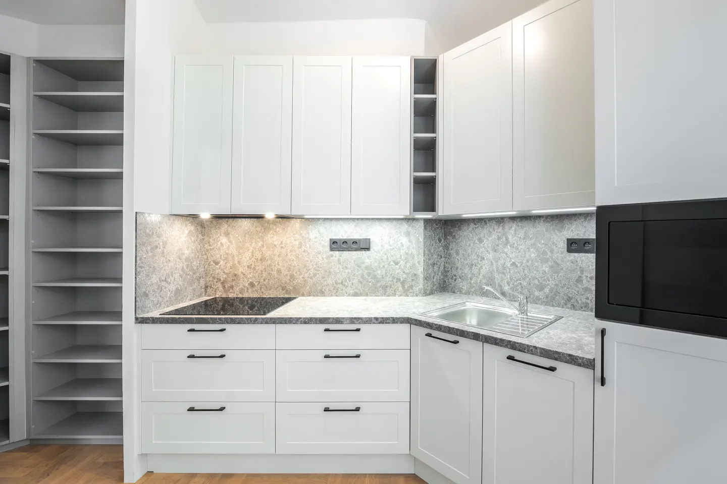 Bright kitchen with white cabinets, black hardware, and gray marble backsplash. Stainless steel sink and black microwave. Open shelving unit on the left.