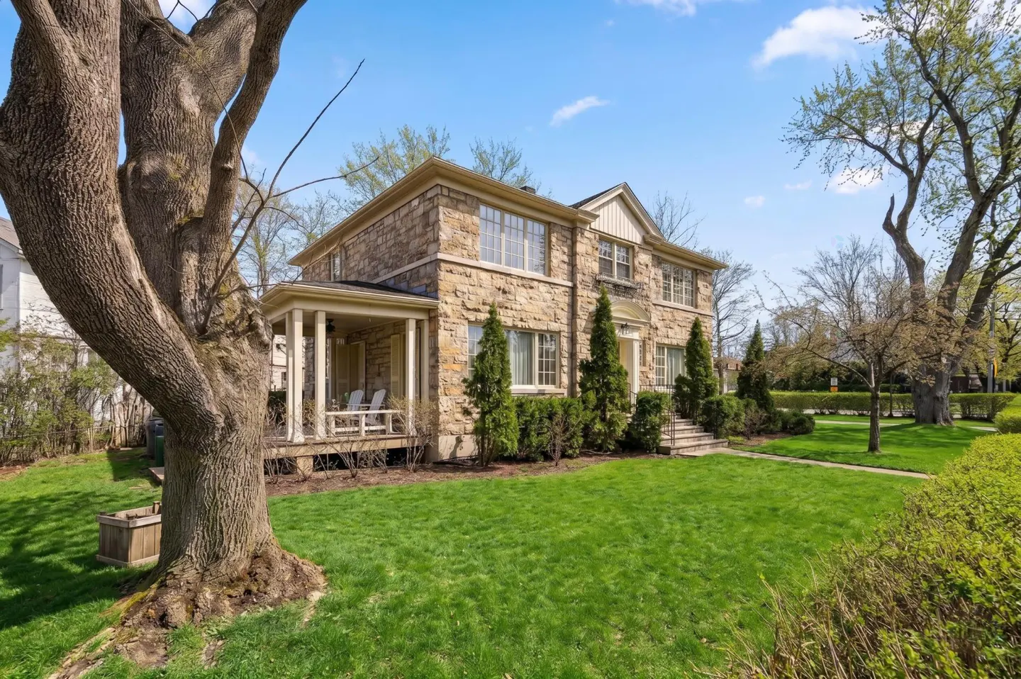 Two-story stone house with a porch, green lawn, and trees under a blue sky.
