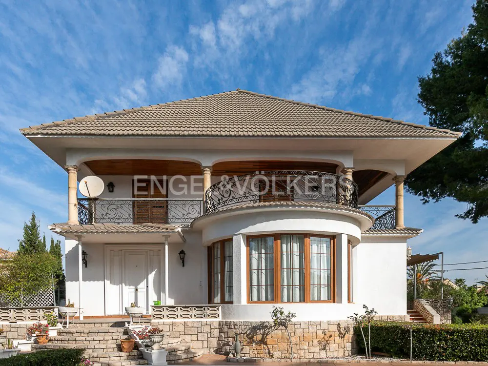 Two-story white house with a brown tile roof, stone base, and wrought iron balcony under a blue sky.