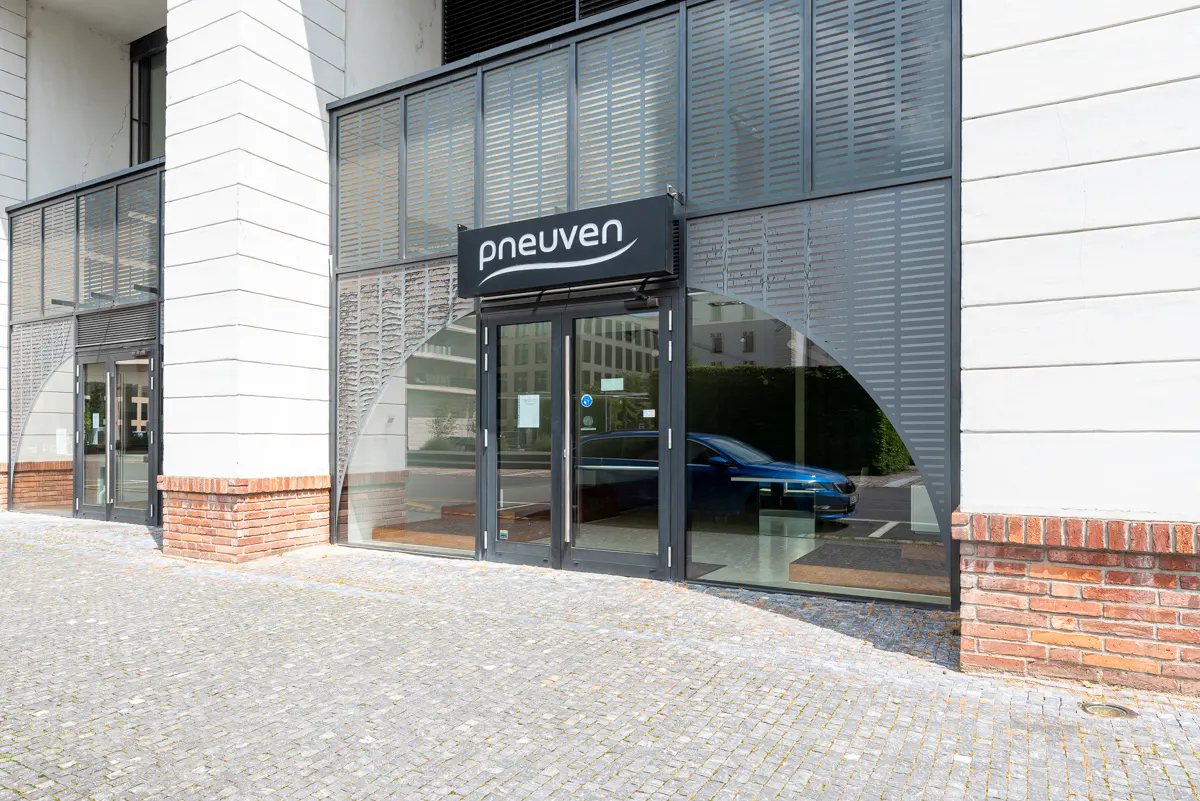 Exterior of "pneuven" building with glass doors and windows, gray metal screens, and a blue car visible inside.