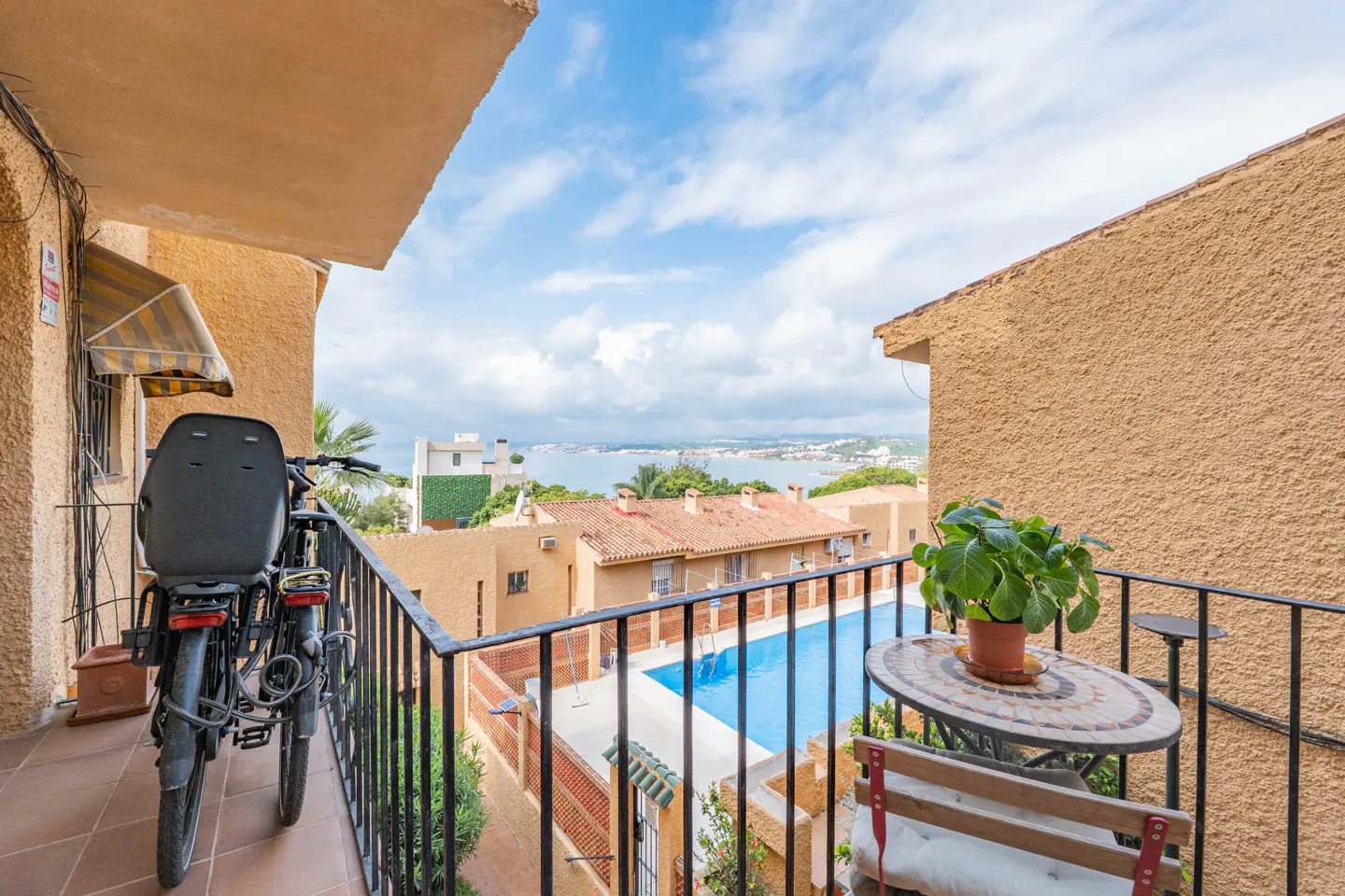 Balcony view with a bicycle, table, and potted plant overlooking a pool and distant ocean under a partly cloudy sky.