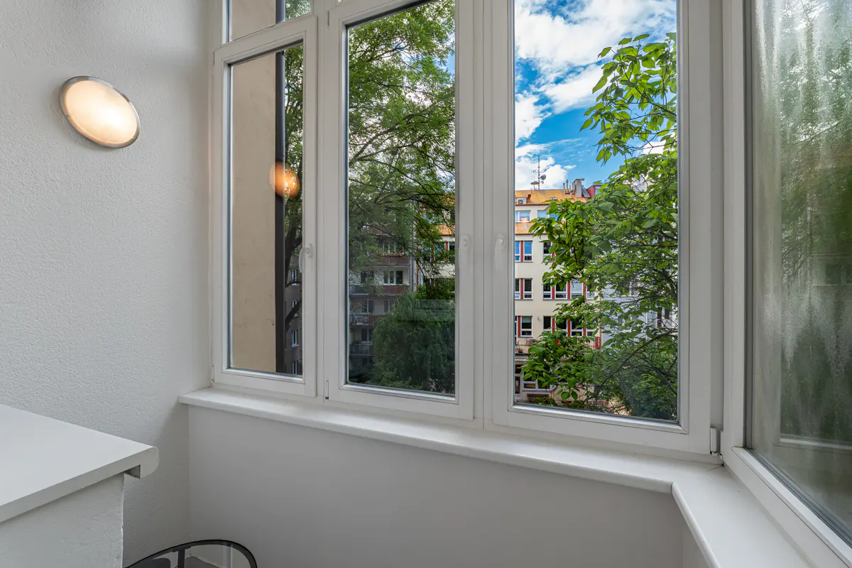 White window frames look out onto green trees and a neighboring apartment building under a blue sky with white clouds.