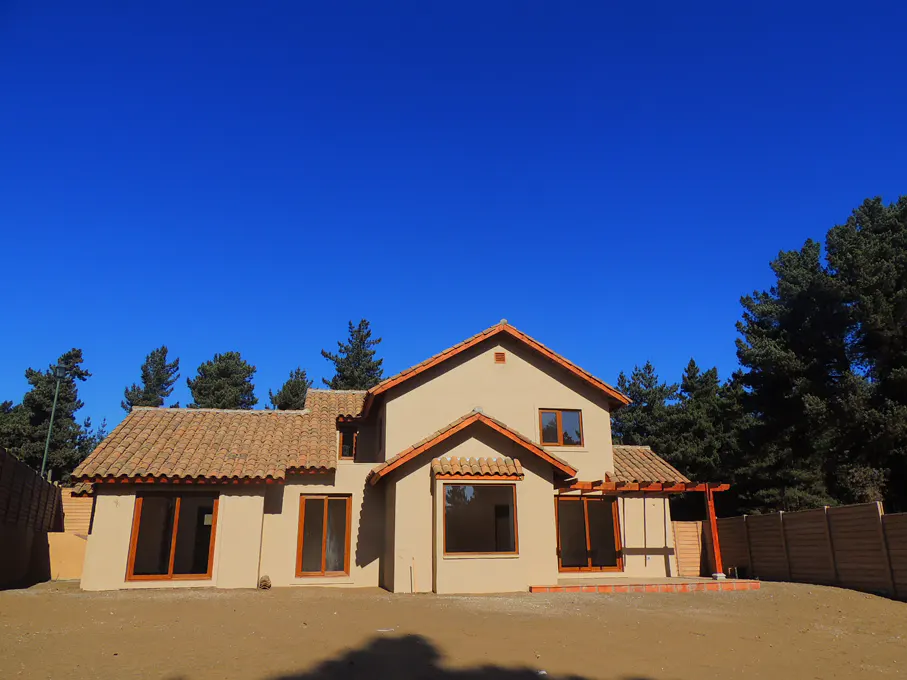 Two-story beige house with a red tile roof, brown trim, and a clear blue sky. Trees are visible in the background.