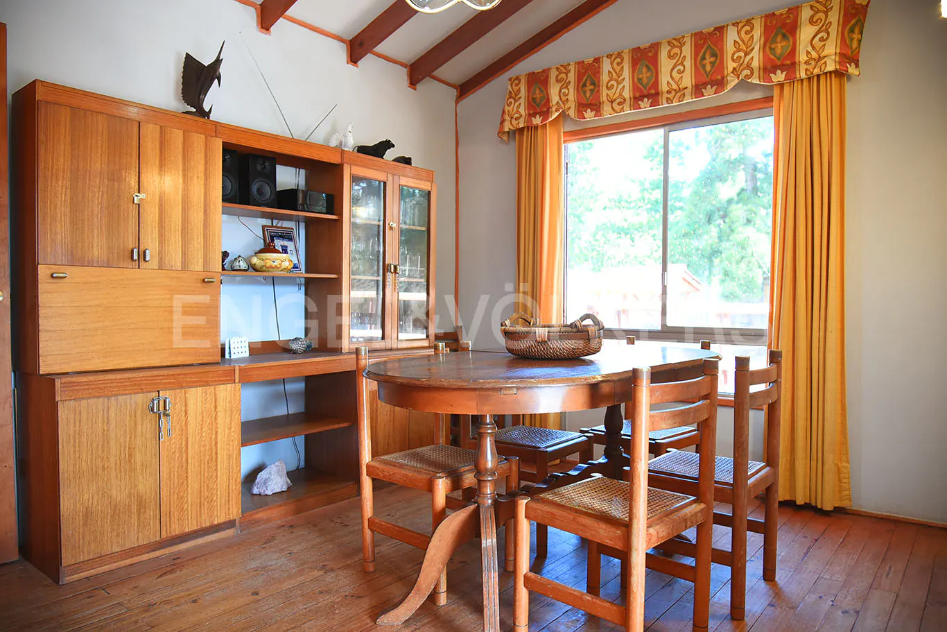 A dining room with a wooden table, chairs, and a large wooden cabinet. A window with yellow curtains is in the background.
