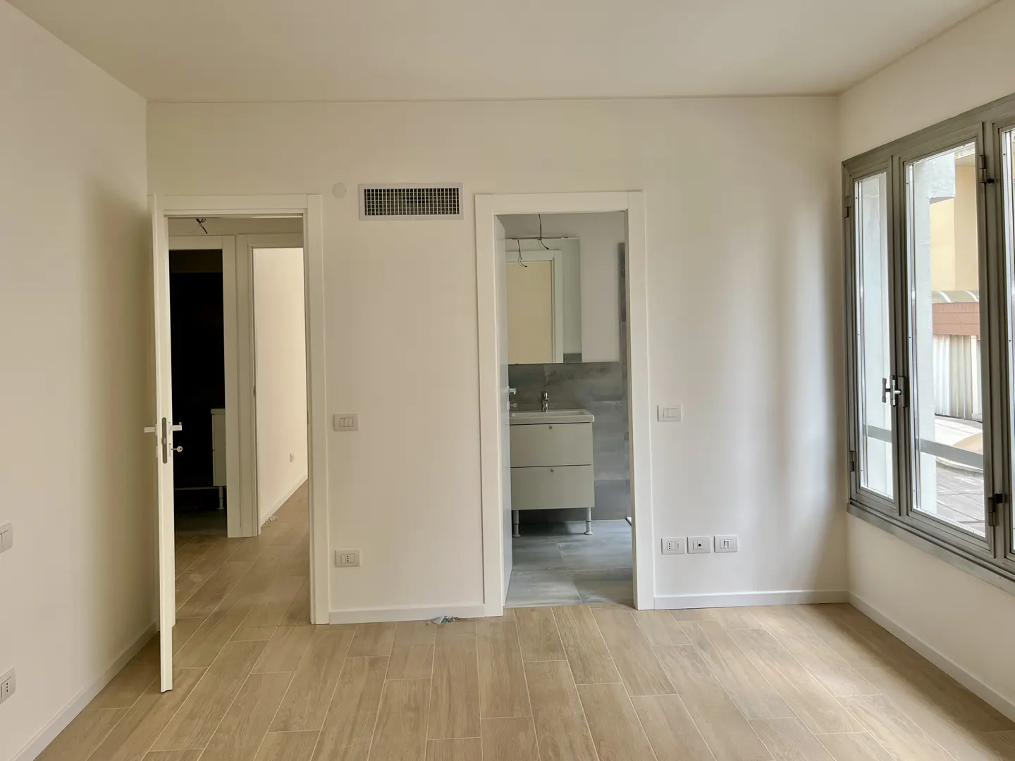 Bright, empty room with wood floors, white walls, and gray-framed windows. Two open doorways lead to other rooms, one to a bathroom.