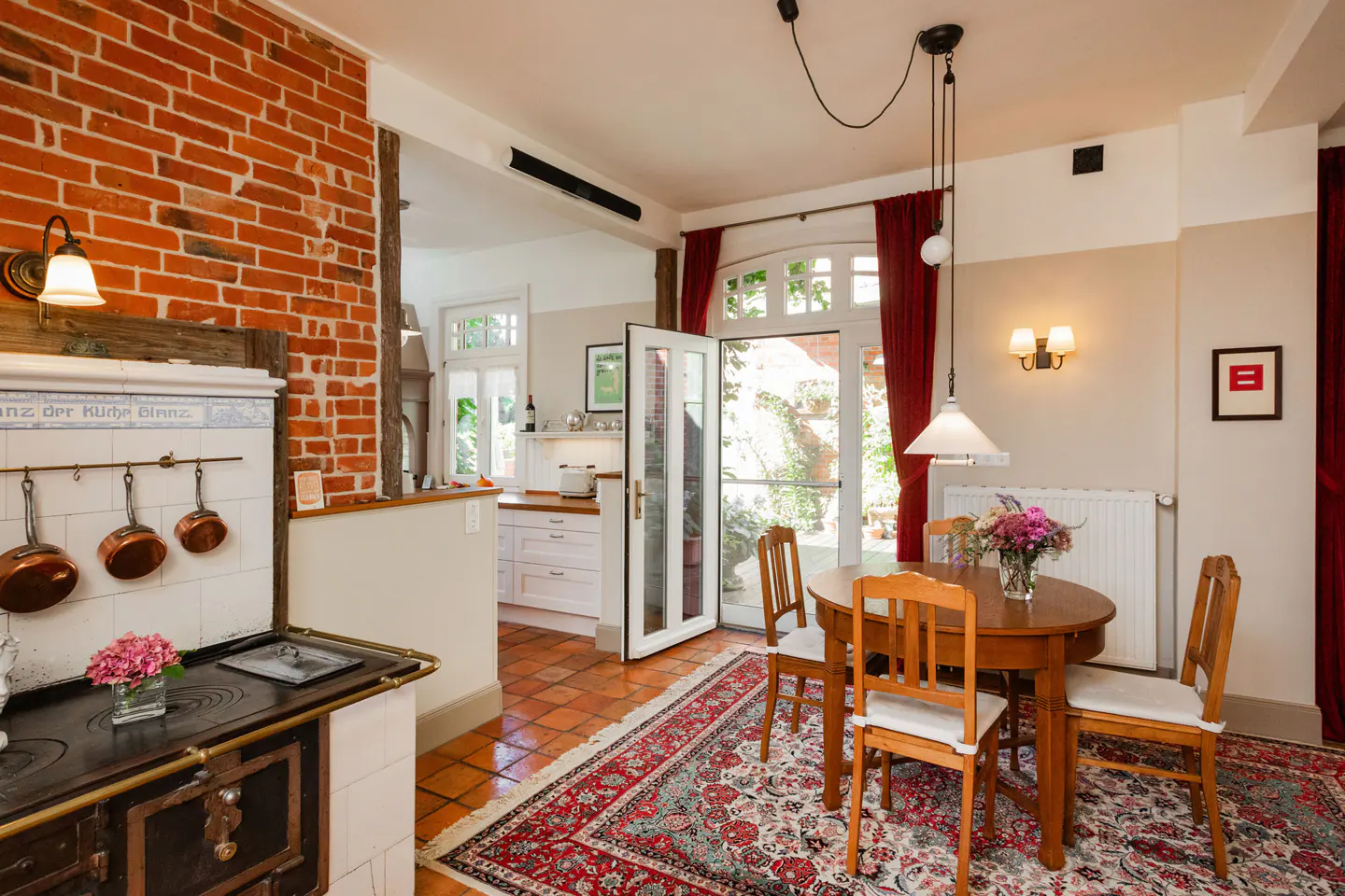 A kitchen and dining area with a brick wall, wood-burning stove, and a round wooden table with chairs. Red curtains frame the open doors.