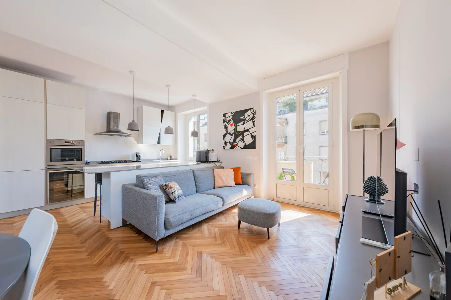 Bright, modern apartment interior with herringbone wood floors, a gray sofa, and a white kitchen with stainless steel appliances. French doors lead to a balcony.