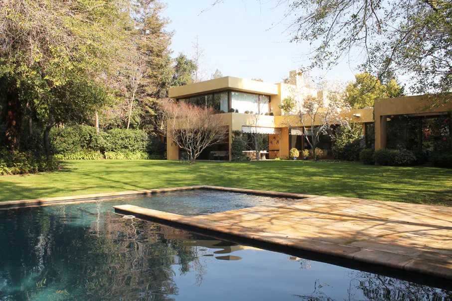 Modern yellow house with large windows overlooks a pool and green lawn surrounded by trees.