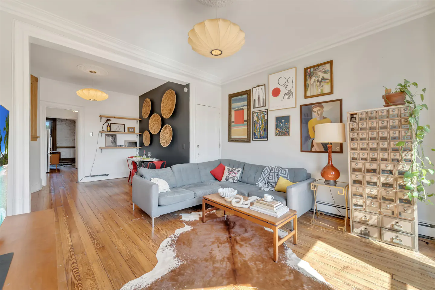 A living room with hardwood floors, a gray sectional sofa, and a cowhide rug. Artwork adorns the walls, and a vintage cabinet stands to the right.