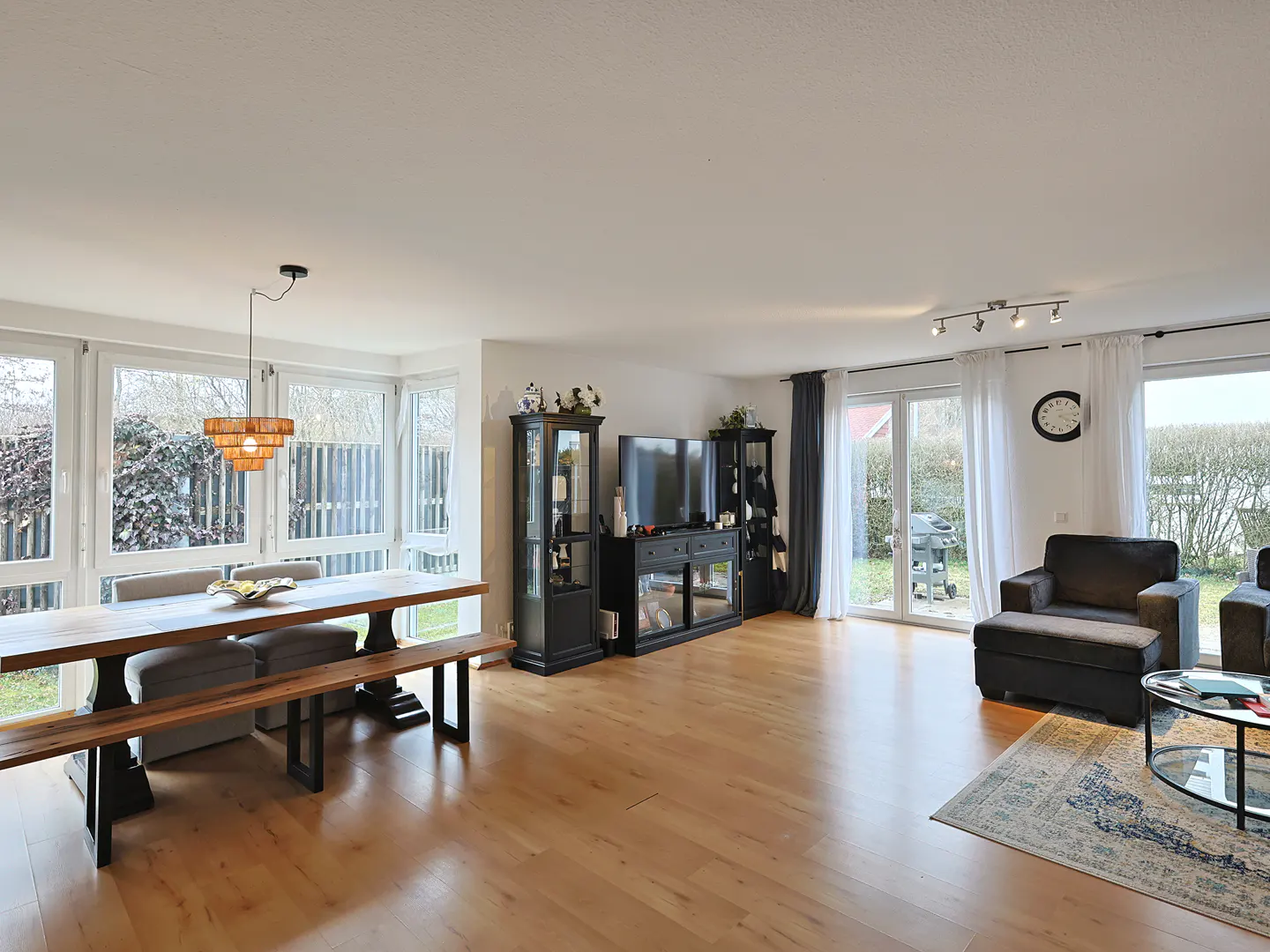 Bright, open-concept living and dining area with wood floors. A large wood table and benches sit near windows. Black cabinets and a TV stand against the wall. Dark sofas and a rug in the living area.