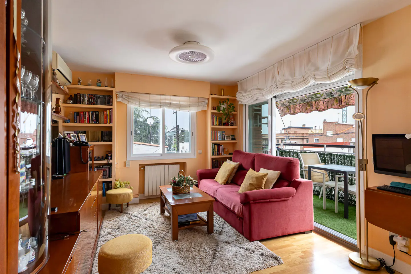 Cozy living room with a red sofa, wooden furniture, bookshelves, and a balcony with city views.