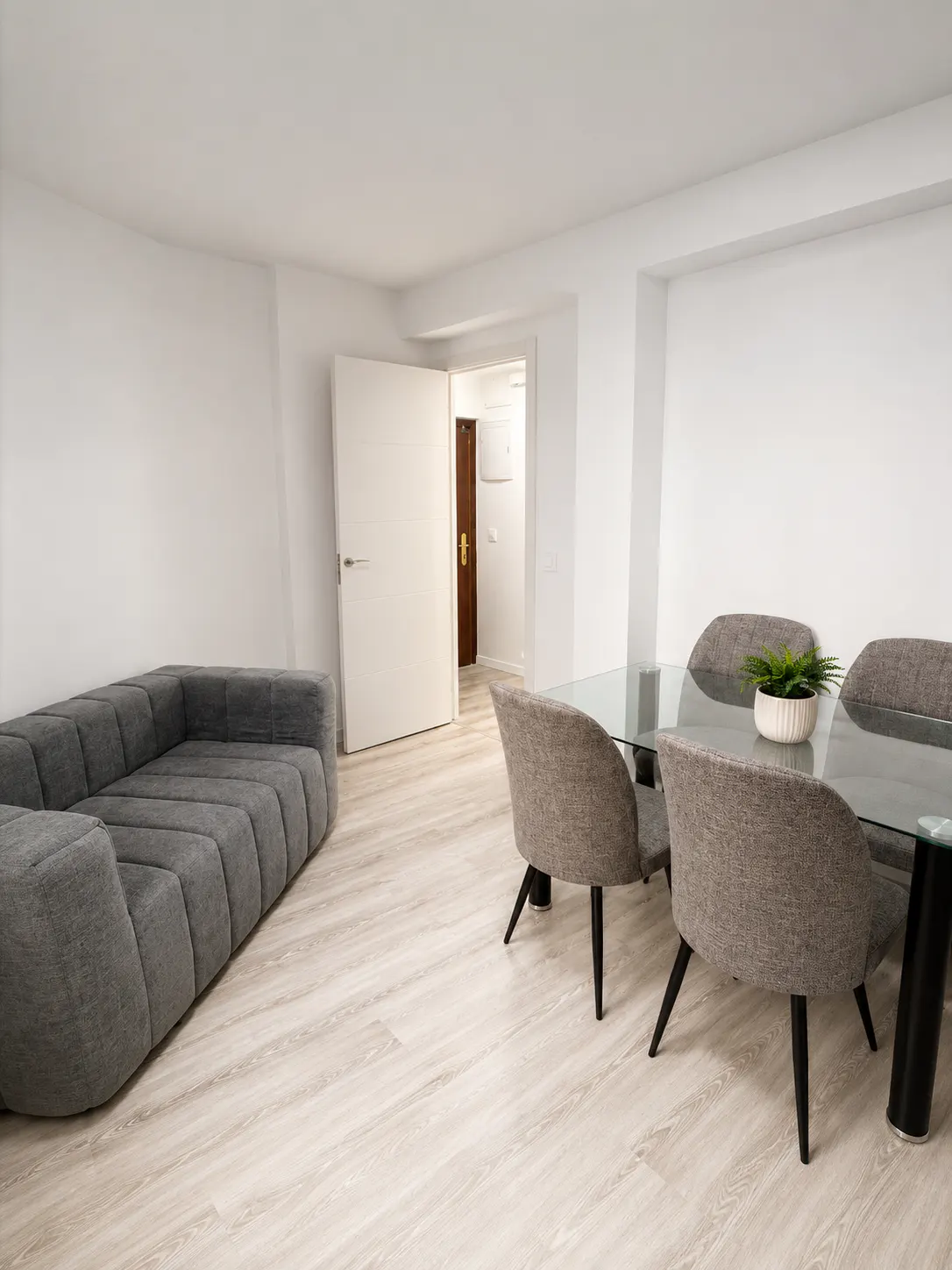 Modern living room with a grey tufted sofa, glass dining table, four grey chairs, and light wood floors.