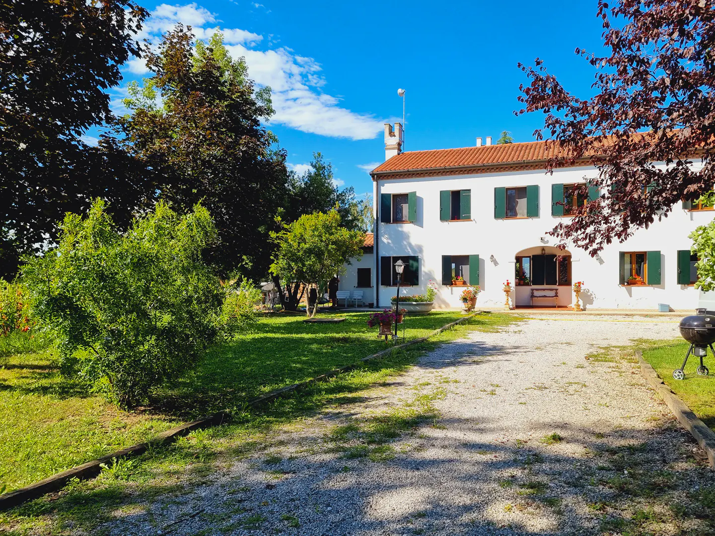 Exterior view of a two-story white house with green shutters, a red tile roof, and a gravel driveway. Trees and lawn surround the house.