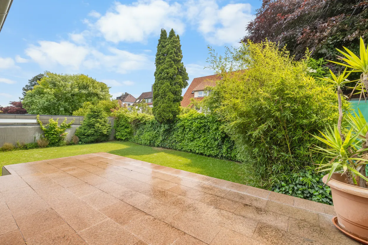 A backyard with a stone patio, green grass, and lush greenery under a blue sky with white clouds.