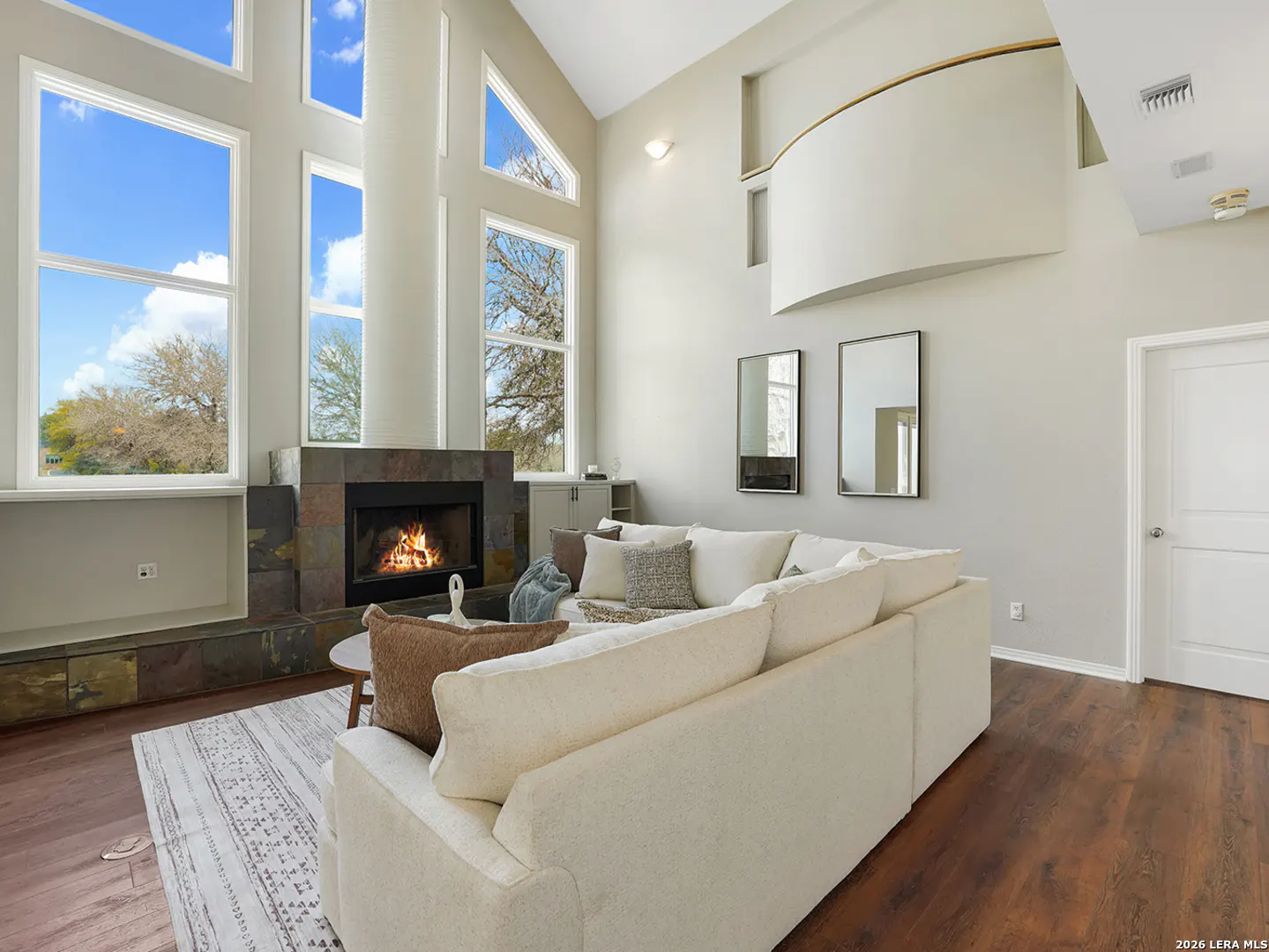 Bright living room with a stone fireplace, large windows, and a cream-colored sectional sofa on a patterned rug.