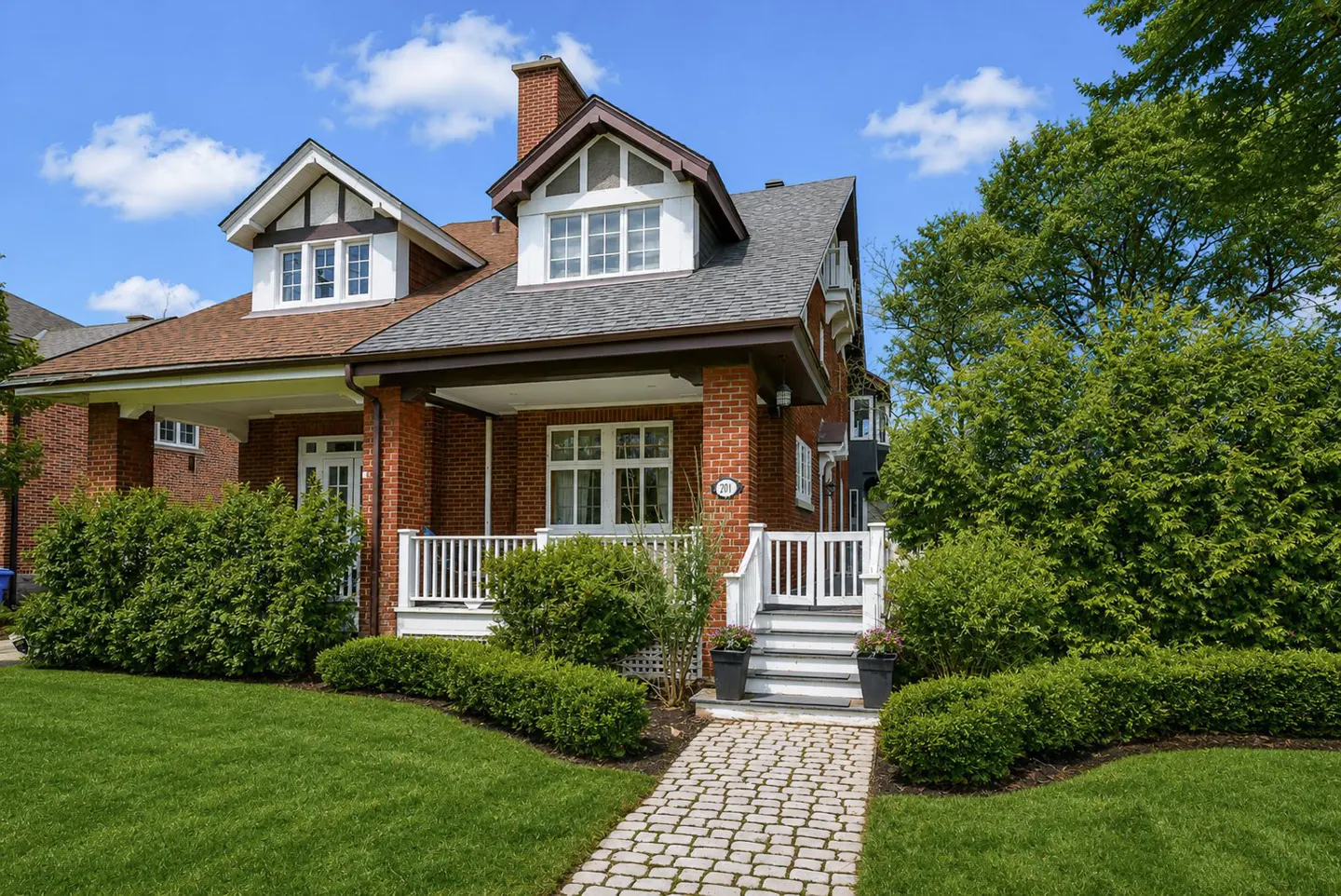 Red brick house with white trim, dormers, and a chimney. A stone path leads to the white porch and gate, surrounded by green lawn and bushes.