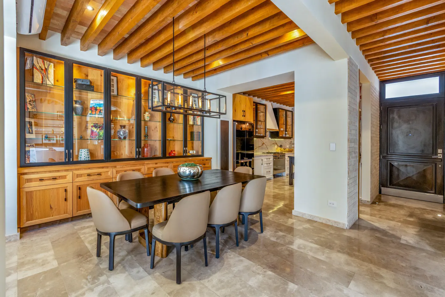 A dining room with a dark wood table, beige chairs, and a wooden ceiling. A glass cabinet displays decorative items. The floor is marble.