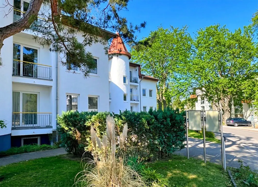 Exterior of a white apartment building with balconies and a red-roofed turret, surrounded by green trees and lawn.