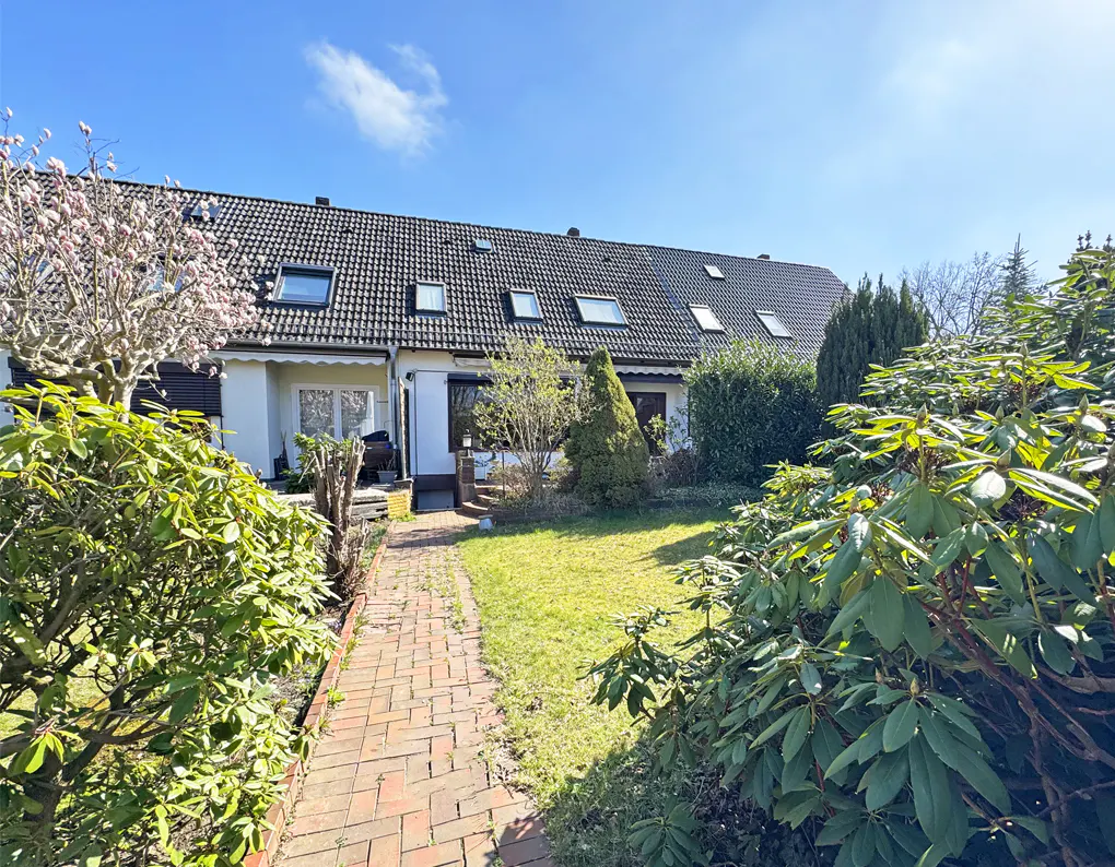 Row of townhouses with gray roofs and white walls, viewed from a brick path through a green lawn and bushes under a blue sky.