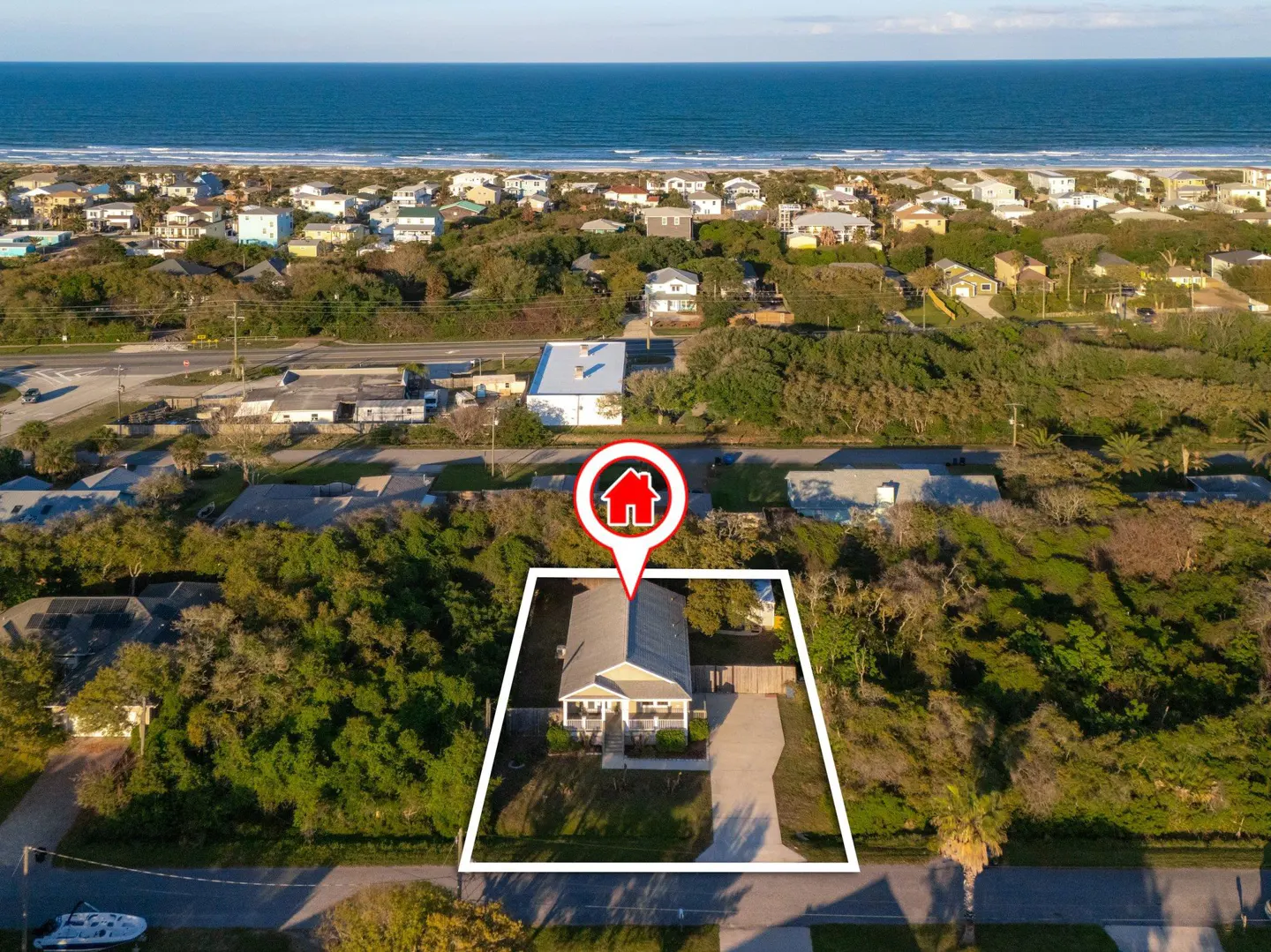 Aerial view of a beige house with a white border, near the beach. A red house icon marks the location. Trees and other houses are visible.