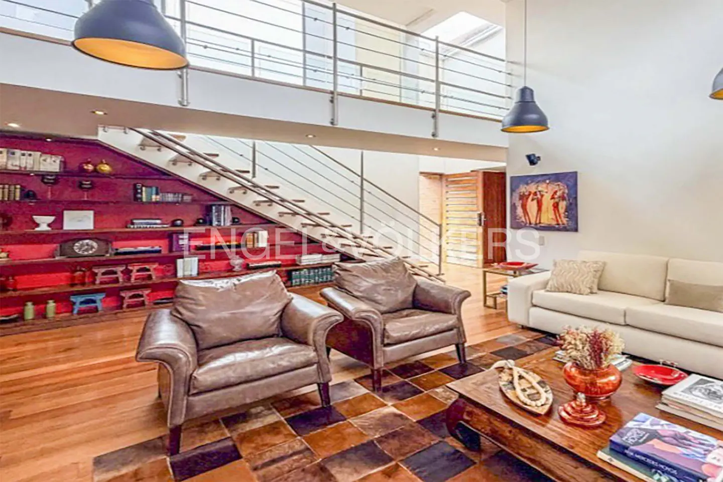 A living room with brown leather chairs, a white sofa, and a wooden coffee table on a brown tiled floor. A staircase leads to a second floor with a metal railing. Red bookshelves are built into the wall.