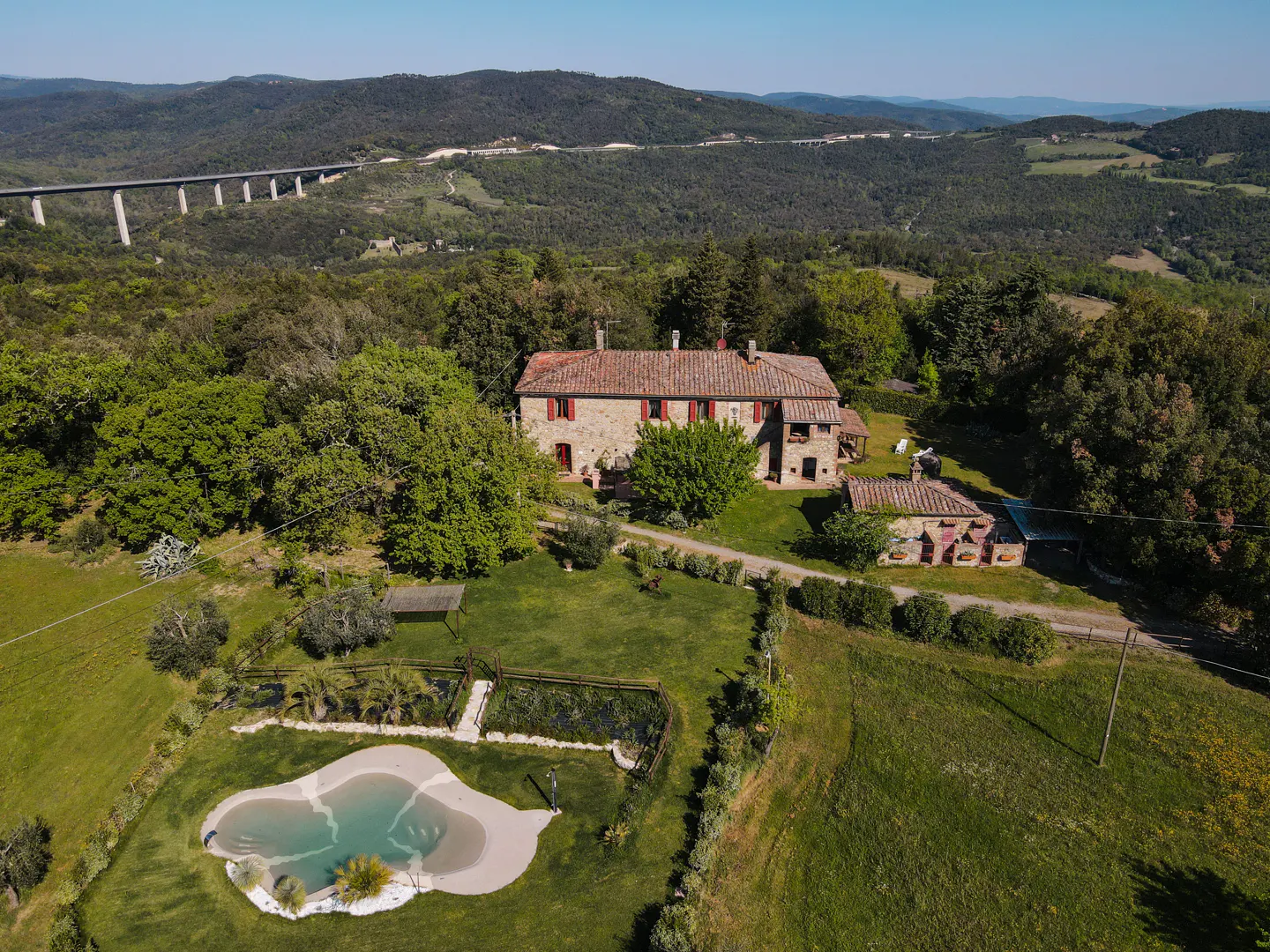 Aerial view of a stone farmhouse with red shutters, a pool, and a bridge in the background.