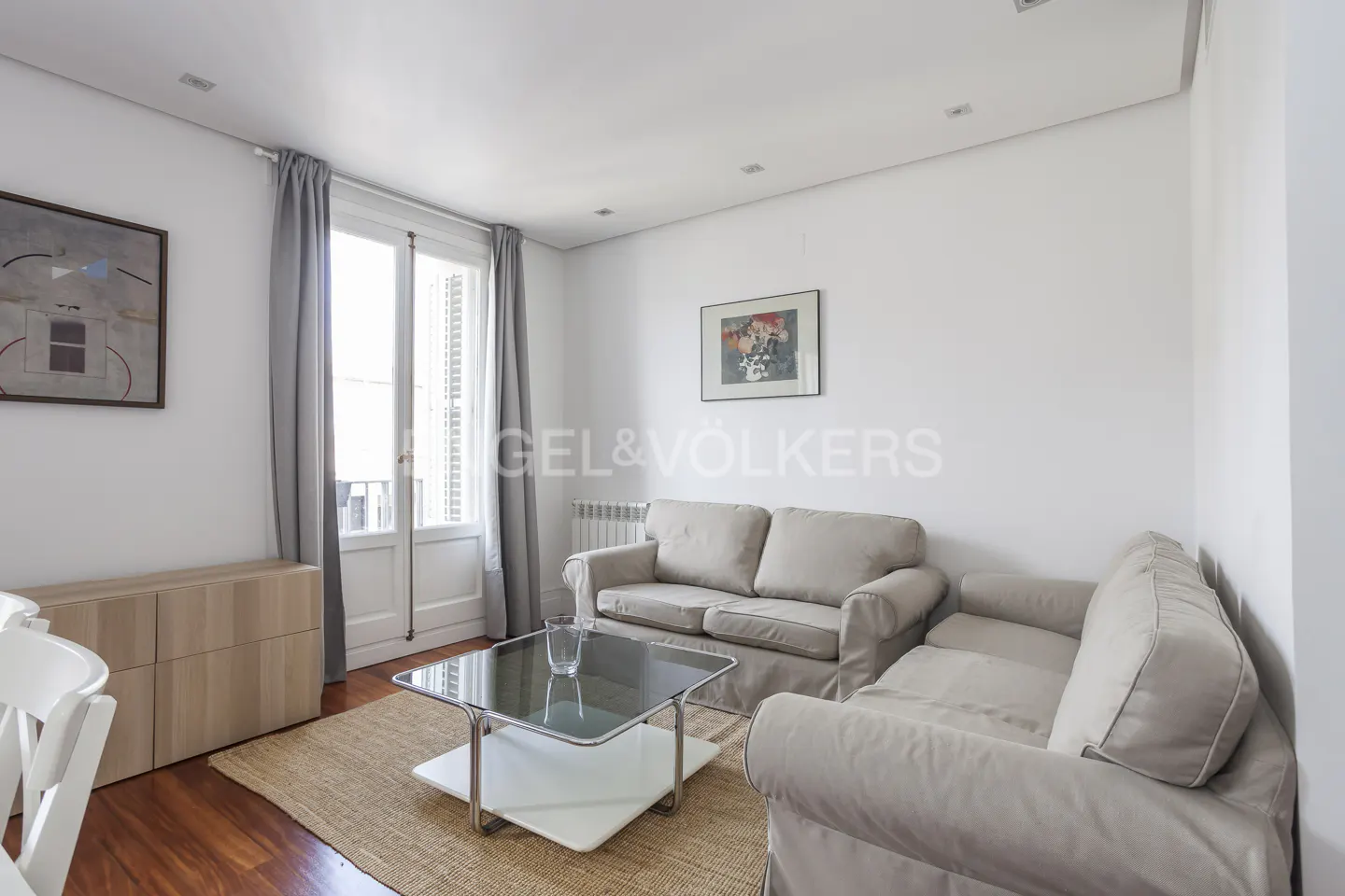 Bright living room with two beige sofas, a glass coffee table, a jute rug, and a balcony door with grey curtains.