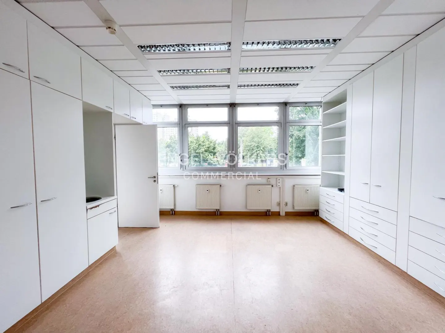 Bright office space with white cabinets, large windows, and a light brown floor. Radiators are under the windows.