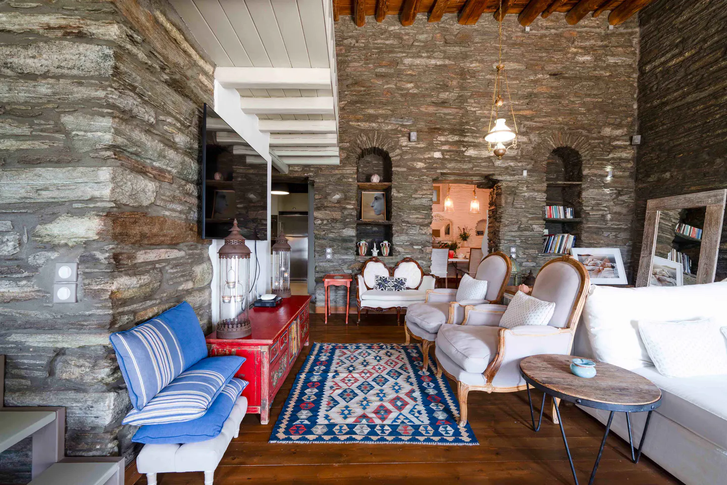 Stone-walled living room with hardwood floors, blue rug, and neutral-toned furniture. A red cabinet adds a pop of color.