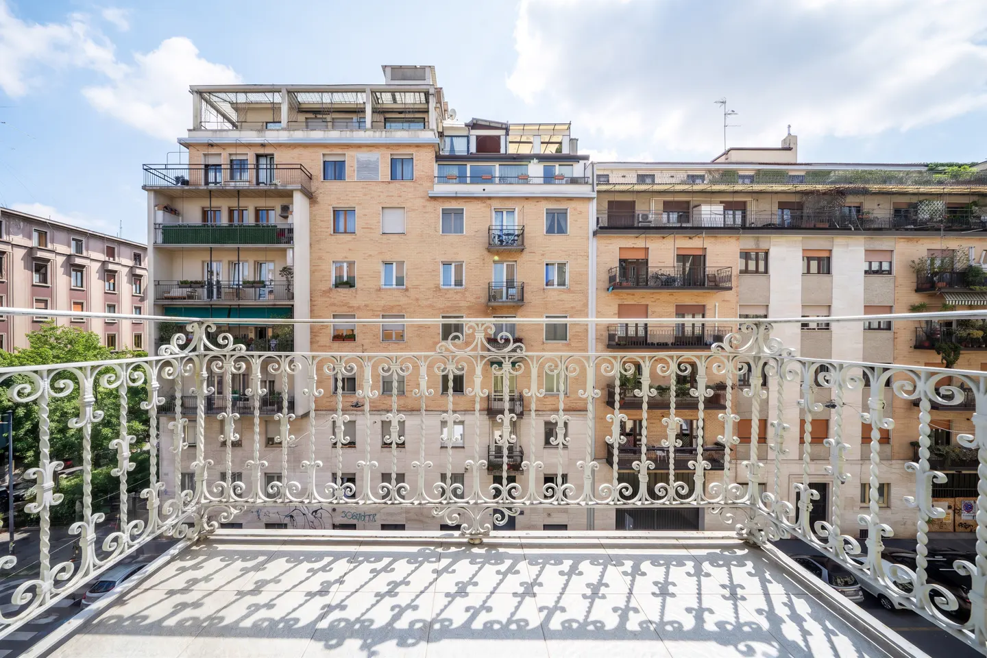 View from a balcony with a white ornate railing, overlooking city buildings under a blue sky with clouds.