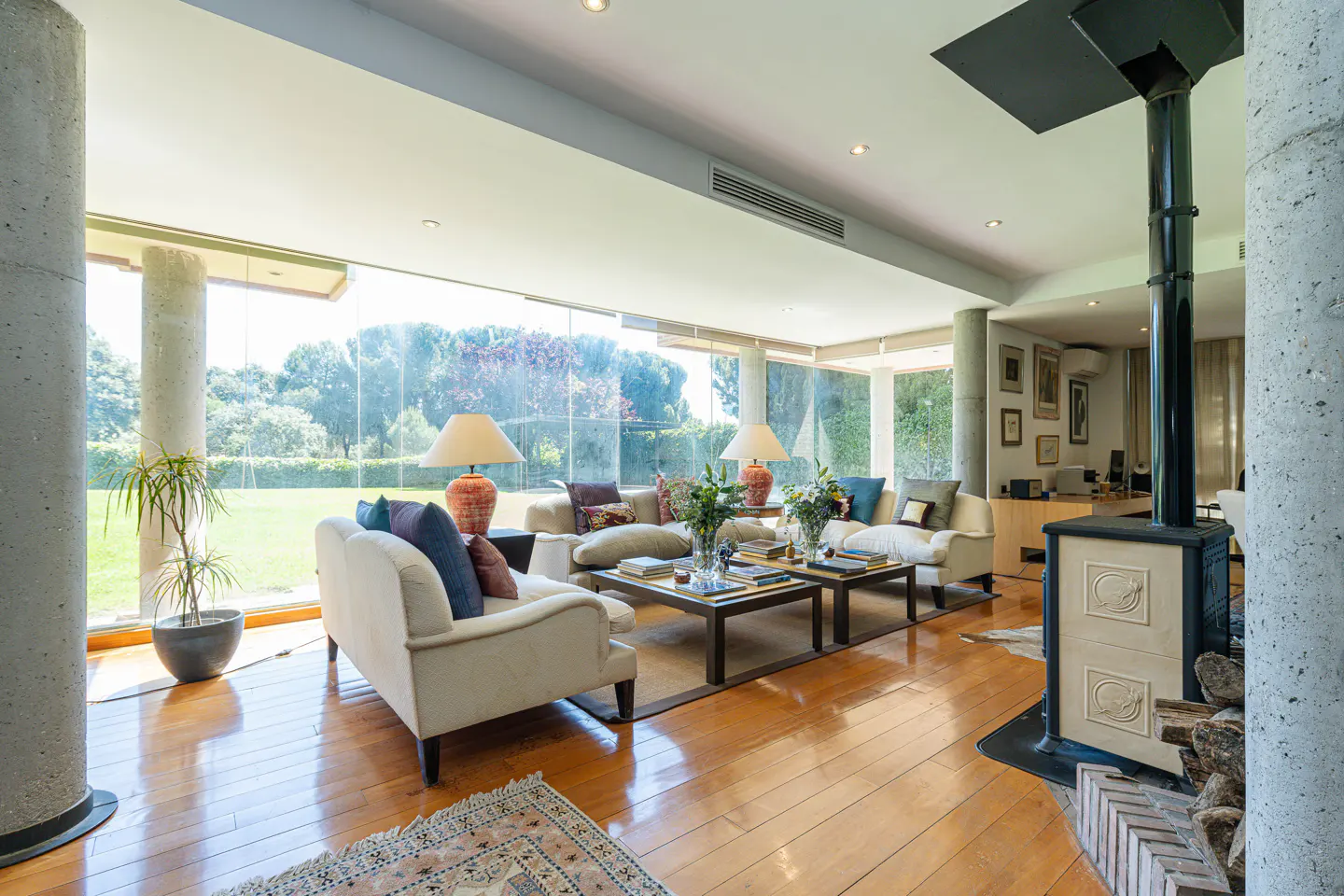 Bright living room with hardwood floors, white sofas, and a modern fireplace. Large windows offer a view of a green lawn.