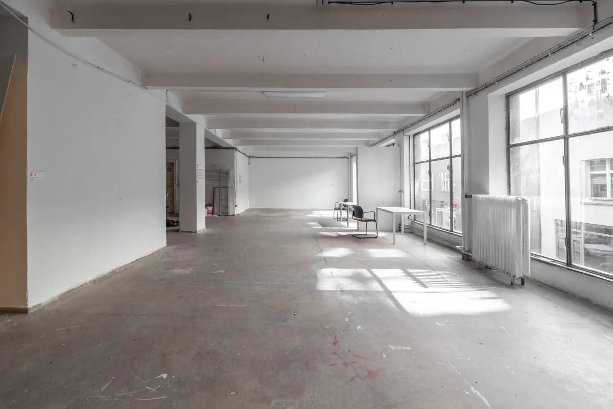 Bright, empty commercial space with white walls, concrete floors, and large windows. A radiator and minimal furniture are present.