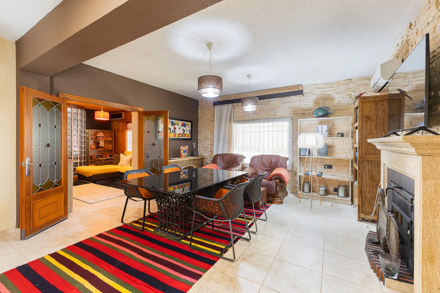 A dining room with a black table, woven chairs, and a colorful striped rug. A fireplace and shelving unit are on the right.