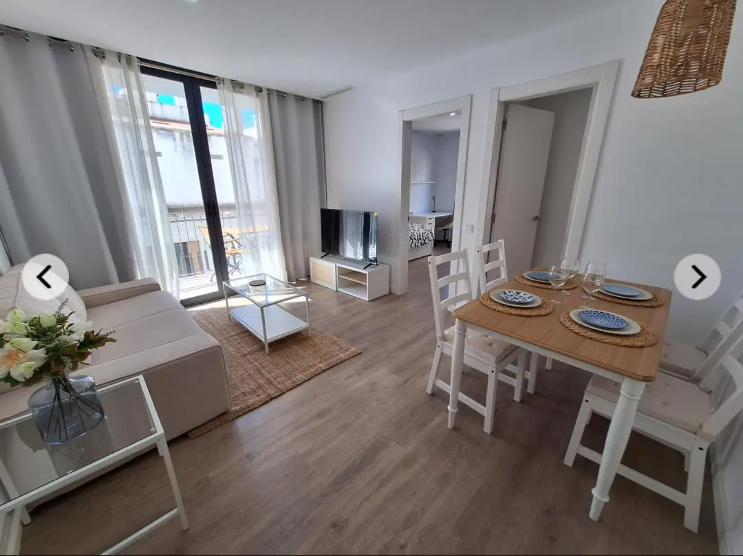 Bright apartment interior with a beige sofa, glass coffee table, TV, and a dining table set for four. Balcony visible through sheer curtains.