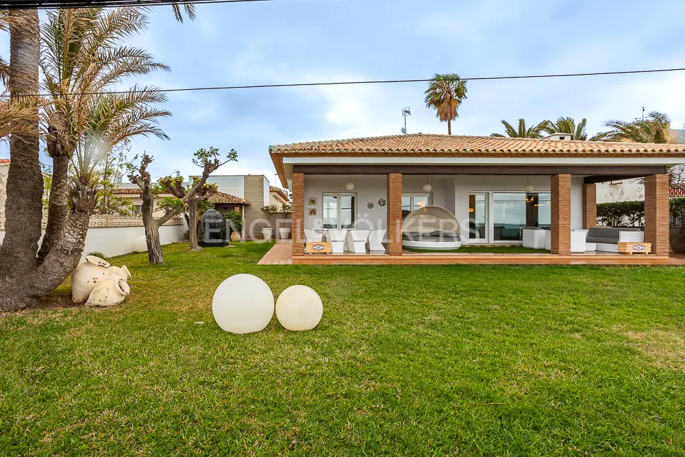 A single-story white house with a red tile roof and brick columns, surrounded by a green lawn with palm trees and decorative spheres.