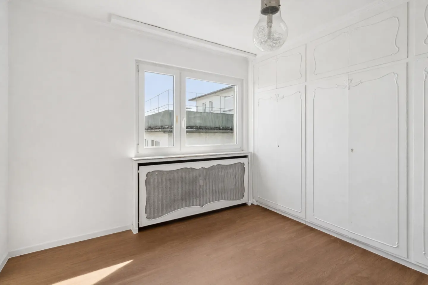 Bright, empty room with wood floor, white walls, window, radiator cover, and built-in white wardrobe.