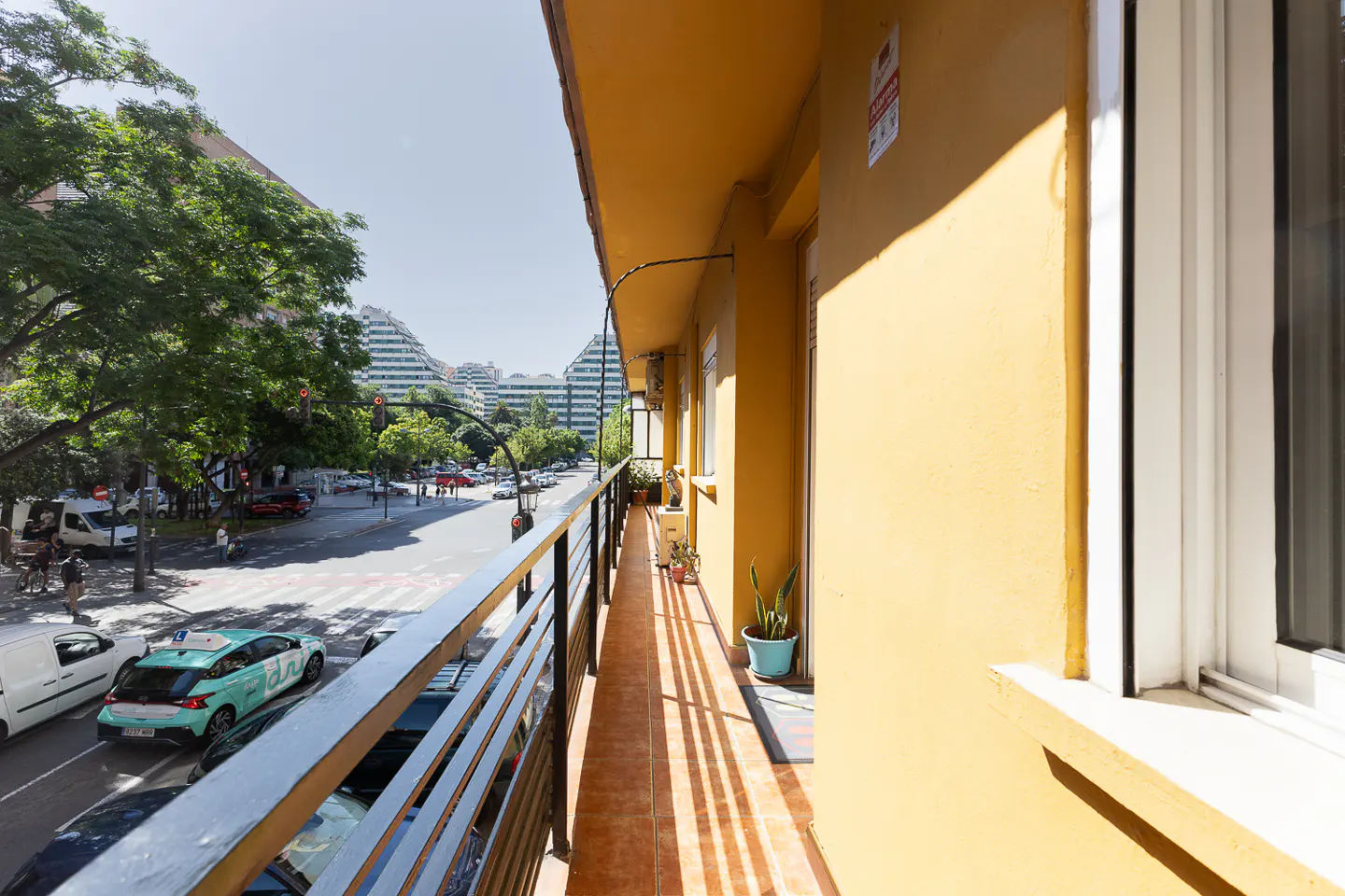 A narrow balcony with orange tiled floors and yellow walls overlooking a busy city street with cars and trees.